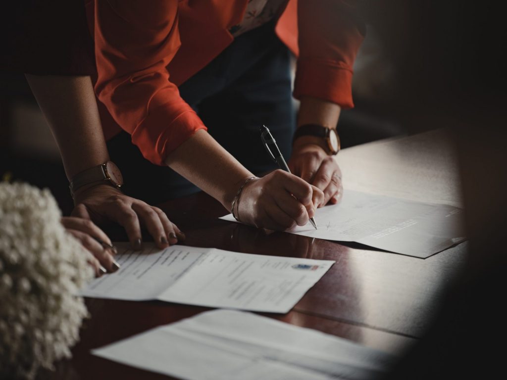 Photo of a person's hand holding a pen to sign contracts placed on a wooden table. - How to Create a Strong Sales Presentation that Drives Conversion