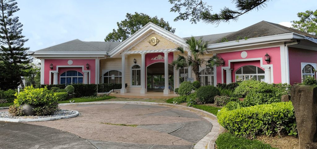 Pink Southern American style club house by a paved rotunda with healthy green pine trees, palm trees, and bushes - Luxury Homes by Brittany Corporation