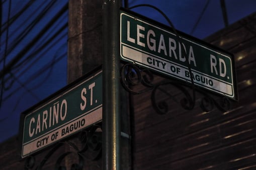 Perpendicularly oriented street name signs at the Cariño-Legarda road junction sparsely illuminated at night by the street lights around. Baguio-Benguet prov.-Cordillera reg.-Luzon island-Philippines