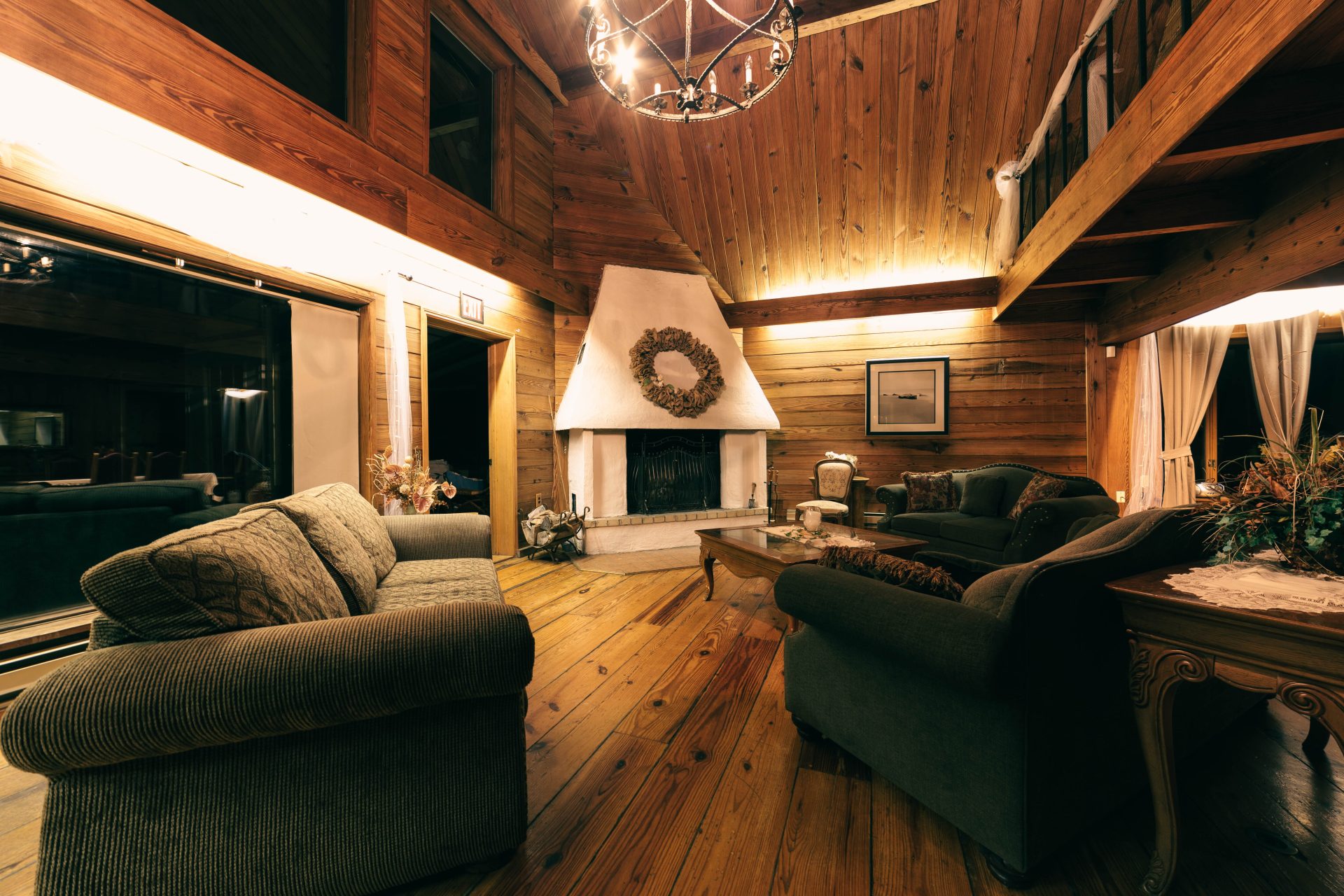 Interior of a wooden house. Includes a central fireplace, expansive windows, and emerald-colored couches.